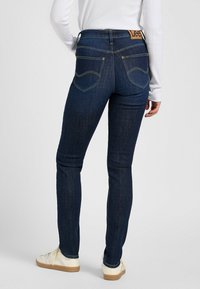 Mörkblå denimjeans med en slät textur, med en läderetikett i midjan och rundade bakfickor med gula stygn.