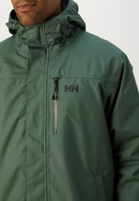 Chaqueta impermeable verde con cuello alto, capucha ajustable y bolsillo en el pecho con cremallera que presenta un logo negro. Textura suave, diseño moderno.