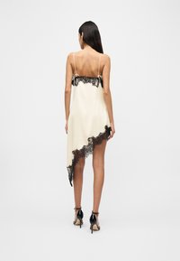 Γυναίκα με μακριά σκούρα μαλλιά φοράει λιτή σατέν κρεμ φόρεμα με μαύρη δαντέλα στο τελείωμα και ασύμμετρο τελείωμα, συνδυασμένο με μαύρα σανδάλια ψηλοτάκουνα.