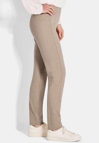 Vrouw in beige slim-fit broek met zijzakken en witte sneakers, zijwaarts staand met één hand op haar heup.