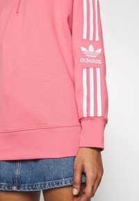 Růžová mikina s vysokým límcem, bílým logem Adidas na rukávu a třemi bílými pruhy podél jednoho rukávu. Měkká tkanina a žebrované manžety.