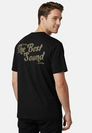 Jonge man draagt een zwart T-shirt met de tekst "The Best Sound" op de achterkant, kijkt over zijn schouder en lacht.