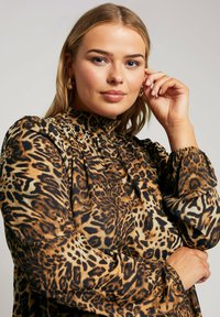 Blusa in stampa leopardata con collare alto, maniche arricciate e una texture setosa nei toni di marrone e nero. Dettagli del motivo ben visibili.