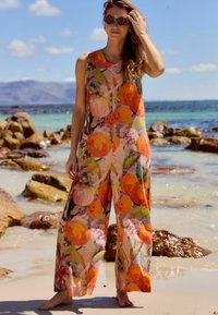 Luftig, ärmlös jumpsuit i ett lätt tyg med ett livfullt orange blommönster på en persikofärgad bakgrund; design med vida ben. Sandstrandsmiljö.