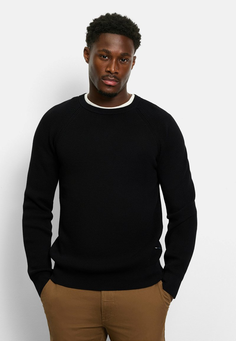 Schwarzer Waffle-Strickpullover mit Rundhalsausschnitt und gerippten Bündchen, kombiniert mit beigen Hosen. Der Pullover zeichnet sich durch den strukturierten Stoff und die enganliegende Passform aus.