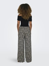 Žena so kučeravými vlasmi stojí chrbtom, oblečená v čiernom topu s odhalenými ramenami a širokých nohaviciach s geometrickými vzormi.