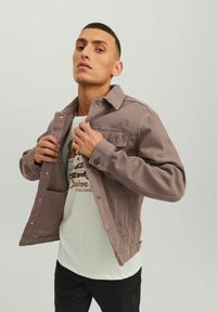 Giacca di denim mauve chiaro con chiusura a bottoni, due tasche sul petto e vestibilità comoda, indossata sopra una t-shirt bianca con stampa e pantaloni neri.