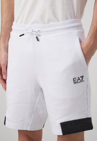Pantalones cortos de algodón blancos con cintura elástica, cordón ajustable, bolsillos laterales y acentos negros en el dobladillo con detalles de logo.