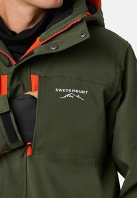 Grüne wasserdichte Jacke mit orangefarbenen Akzenten, ausgestattet mit einer Reißverschlusstasche, verstellbarem Kapuze und Logodetails auf der Brust. Weiche Textur.