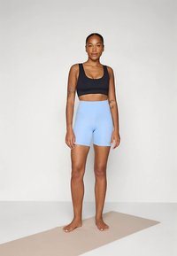 Črni športni nedrček in svetlo modre kratke hlače z visokim pasom, obe iz raztegljivega materiala. Kratki lasje, stoji na bež jogijski blazini.