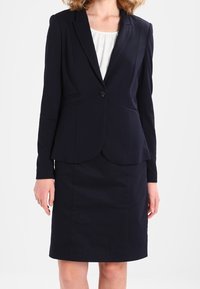 Kvinna klädd i en marinblå kavaj med en enda knapp och matchande knälång pennkjol över en vit blus.