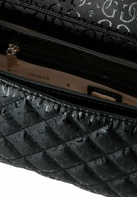 Sac matelassé noir avec un motif de logo brillant, présentant une poche zippée ouverte montrant une doublure intérieure beige avec le texte de la marque "GUESS".