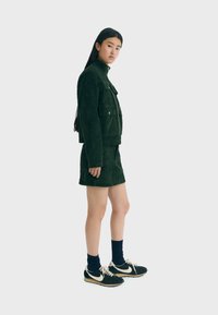 Chaqueta de pana verde con dos bolsillos frontales, combinada con una falda corta de pana y zapatillas negras con detalles y cordones blancos.