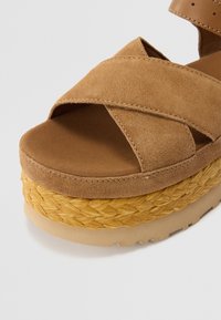 Sandalias de plataforma de ante marrón con tiras cruzadas, media suela de yute trenzado y suela beige texturizada. Diseño limpio, apariencia ligera.