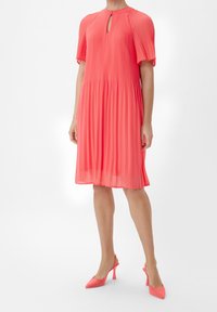 Robe coral plissée avec un décolleté en trou de serrure, des manches courtes et une silhouette évasée. Associée à des talons slingback coral assortis.