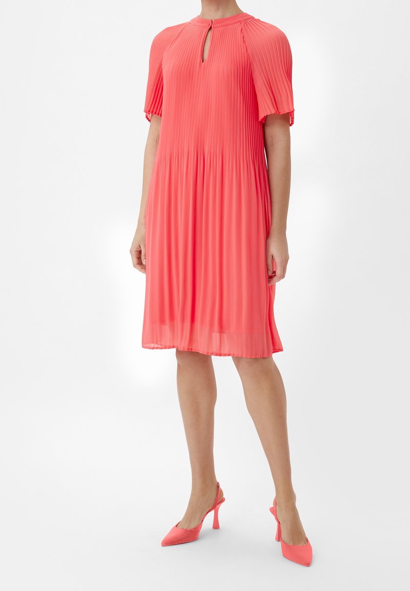 Robe coral plissée avec un décolleté en trou de serrure, des manches courtes et une silhouette évasée. Associée à des talons slingback coral assortis.