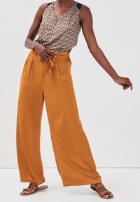 Bedrucktes ärmelloses Oberteil mit V-Ausschnitt und schwarzen Trägern, kombiniert mit weiten, hoch taillierten Hosen in Senfgelb-Orange. Braune Rutsch-Sandalen.