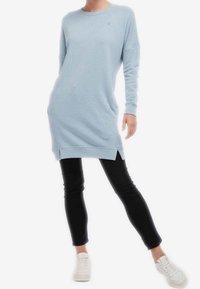 Abito felpa azzurro chiaro con maniche lunghe, scollatura tonda, fessure laterali e un discreto dettaglio con logo. Abbinato a pantaloni neri aderenti e scarpe bianche.