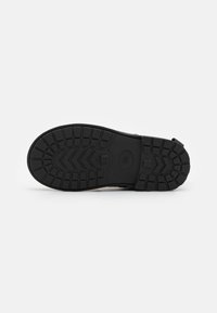 Semelle de chaussure en caoutchouc noir avec un motif de grip texturé. Présente un design de bande de roulement hexagonal et un détail de marque moulé. Forme plate et rectangulaire.