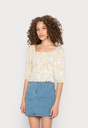 Blusa de padrão floral, em tons pastel, com mangas bufantes e decote quadrado. Combinada com uma saia de ganga clara com costuras visíveis.