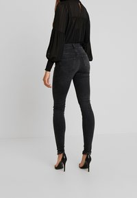 Jeans ajustados de talle alto en negro con un acabado suave, que incluyen bolsillos traseros, combinados con una blusa negra transparente y zapatos de tacón.