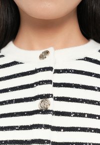 Weißer Cardigan mit navyblauen horizontalen Streifen, verziert mit paillettenbesetzten Akzenten und einer strukturierten Oberfläche, geschlossen mit einem dekorativen silbernen Knopf.