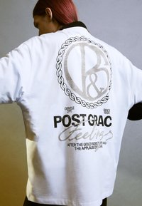 T-shirt bianco oversize con colletto nero. Presenta una grande stampa grafica in argento e nero sul retro, compreso testo e un design circolare.