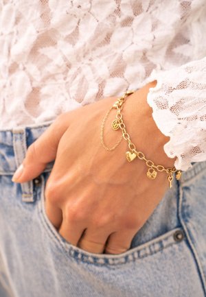 Main portant un bracelet à breloques en or avec des pendentifs en forme de cœur, de serrure et de clé, reposant sur une poche de jean en denim bleu clair et une manche en dentelle blanche.