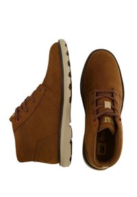 Botas de tobillo de ante marrón con una suela de goma beige, puntera redonda, diseño con cordones y un discreto logotipo en la lengüeta. Paneles laterales texturizados.