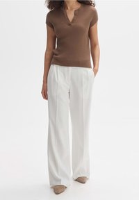 Kobieta w brązowej koszulce z krótkim rękawem w prążki, białych szerokich spodniach i beżowych czółenkach, stojąca z jedną ręką w kieszeni.