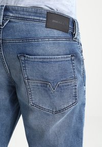 Jeans de mezclilla azul con un acabado desgastado, que cuentan con un parche de cuero etiquetado "DIESEL" en la cintura y un diseño de costura de bolsillo en forma de V.