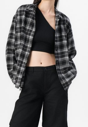 Femme portant un crop top noir à une épaule, une chemise à carreaux noire et blanche oversize, et un pantalon noir taille haute, debout devant un fond blanc.