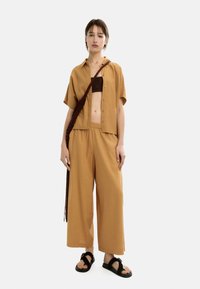 Kobieta stojąca z neutralnym wyrazem twarzy, ubrana w beżową koszulę na guziki, pasujące szerokie spodnie, brązowy bandeau, czarne sandały i brązową torbę na ramię.