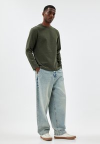 Maglietta a maniche lunghe a coste verde scuro abbinata a jeans oversized in denim azzurro e sneakers bianche con dettagli marroni.