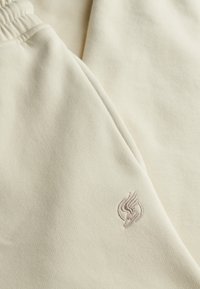 Gros plan sur un tissu couleur crème avec un logo brodé représentant un pied ailé près de la ceinture élastiquée.