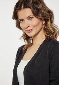 Zwarte cardigan met een V-halsontwerp, lichtgewicht textuur en lange mouwen, gedragen over een witte tanktop. Zacht materiaal, eenvoudige esthetiek.