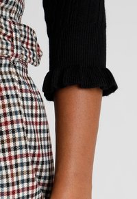 Czarny sweter z prążkowanym rękawem zakończonym falbanką, zestawiony z sukienką w kratę o wzorach czerwonych, brązowych i niebieskich.