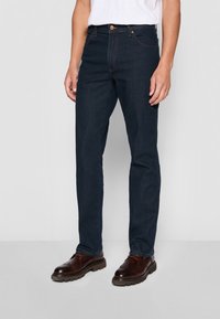 Vaqueros de mezclilla azul oscuro, con un ajuste ceñido, diseño tradicional de cinco bolsillos y costuras marrones, combinados con zapatos marrón oscuro.