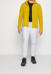 Fleecejacka i klar gul färg, zip-up-design som bärs med svart t-shirt, vita byxor och svarta vandringsskor med snörning.