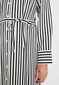 Čierno-biely pruhovaný šaty s gombíkovou prednou časťou, zaväzovaným pásom a dlhými rukávmi s kontrastnými manžetami. Textúrovaná tkanina.