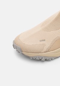 Beige slip-on sko med en åndbar mesh-overside, tekstureret mønster og en fleksibel grå gummisål med trækkraft-studs.