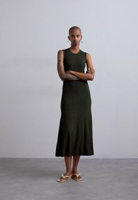 Donkergroene, geribbelde, mouwloze jurk met een rechte silhoutte, die onder de knie valt, gecombineerd met lichtgekleurde platte sandalen.