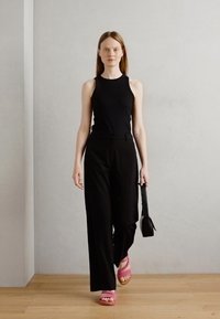 Svart ermeløs topp, brede svarte bukser og rosa sandaler. En liten svart veske holdes i hånden. Glatt, minimalistisk design på en lys bakgrunn.