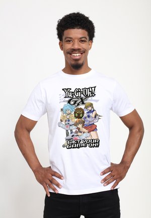 Glunderende man met krullend haar draagt een wit Yu-Gi-Oh GX T-shirt met drie animefiguren en de tekst "Get Your Game On!"