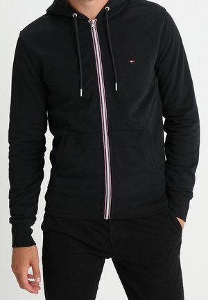 Schwarze Zip-Hoodie mit einem kontrastierenden roten und weißen Streifen entlang des Reißverschlusses. Verfügt über eine Fronttasche und eine Kapuze mit Kordelzug. Glatte Stoffoberfläche.