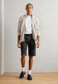 Ljus beige ribbade kavaj, vit t-shirt, svarta denimshorts, svarta slip-on skor, vita strumpor; står mot en ljusgrå vägg.