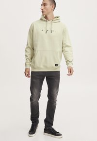 Lys grøn hættetrøje med en frontlomme og snoretræk. Har en sort logo tekst "A.R.I.C." på brystet. Båret med mørkegrå jeans og sorte sneakers.