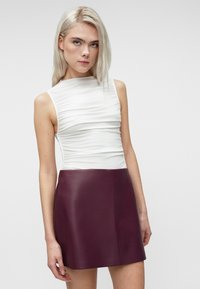 Jonge vrouw met blond haar draagt een gerimpeld wit mouwloos topje en een donkerpaarse leren minirok tegen een effen lichte achtergrond.