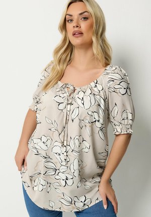 Femme portant un chemisier beige clair avec un motif floral noir et blanc, des manches courtes bouffantes et un col noué devant, associé à un jean bleu.