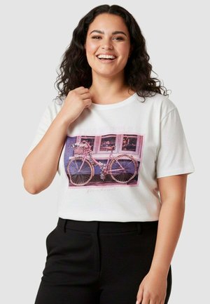 Donna sorridente con capelli ricci scuri indossa una t-shirt bianca con grafica di bicicletta rosa e pantaloni neri, in piedi su sfondo chiaro.